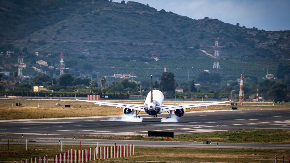 An airplane taking off with smoke from its tires, runway, and surrounding landscape with hills and buildings.