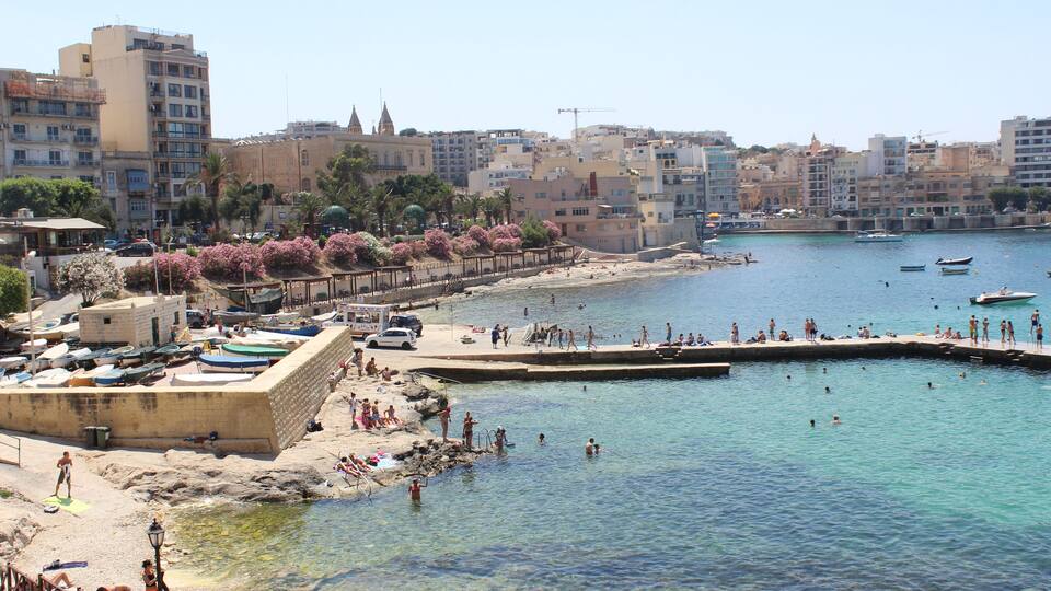 A coastal town with a sandy beach, clear blue water, boats, and buildings in the background.