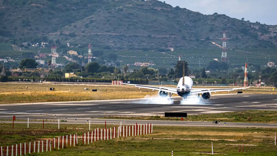 An airplane landing with smoke on a runway, surrounded by grassy fields and a hilly landscape with buildings in the background.