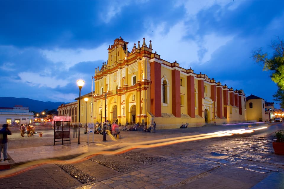 Uma igreja histórica com arquitetura elaborada e um campanário, rodeada por uma praça de calçada e luminárias.