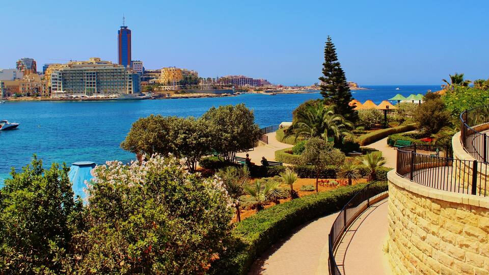 A coastal cityscape with a marina, a tall building, and a landscaped park with trees and a curved stone wall.