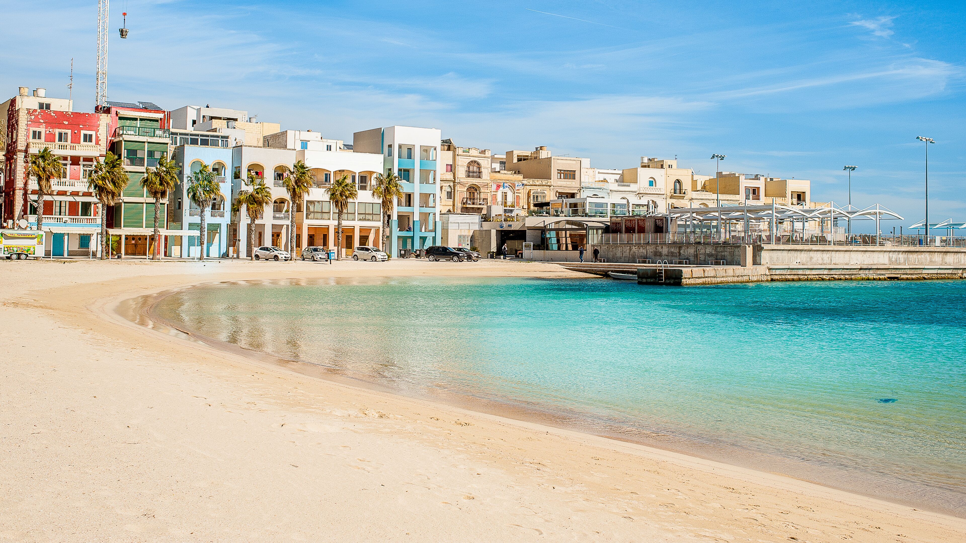A beach with clear turquoise water, a sandy shore, and colorful buildings in the background.