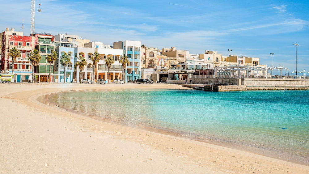 A beach with clear turquoise water, a sandy shore, and colorful buildings in the background.
