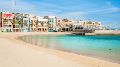 Uma praia com águas turquesa cristalinas, uma orla arenosa e edifícios coloridos ao fundo.