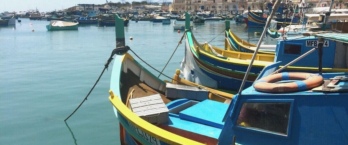 A harbor with several colorful boats docked, a lifebuoy, and buildings in the background.