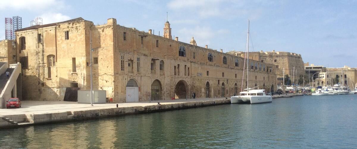 Un bâtiment historique en pierre, doté d’arches au niveau des fenêtres et surmonté d’une tour horlogère, se dresse au bord d’un port tranquille où sont amarrés plusieurs bateaux.