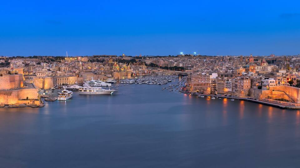 A coastal cityscape with historic buildings, a fortified harbor, and a calm sea at dusk.