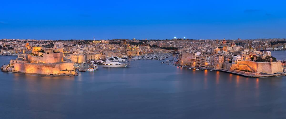 A coastal cityscape with historic buildings, a fortified harbor, and a calm sea at dusk.