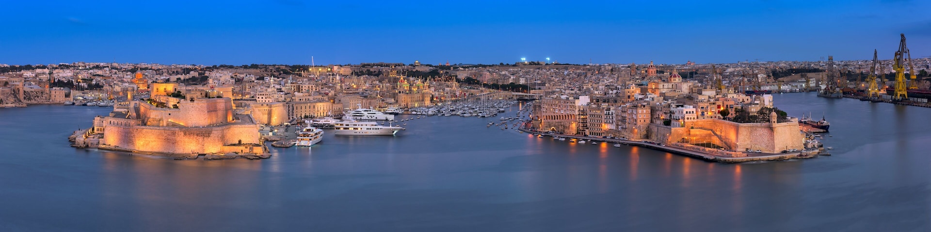 A coastal cityscape with historic buildings, a fortified harbor, and a calm sea at dusk.