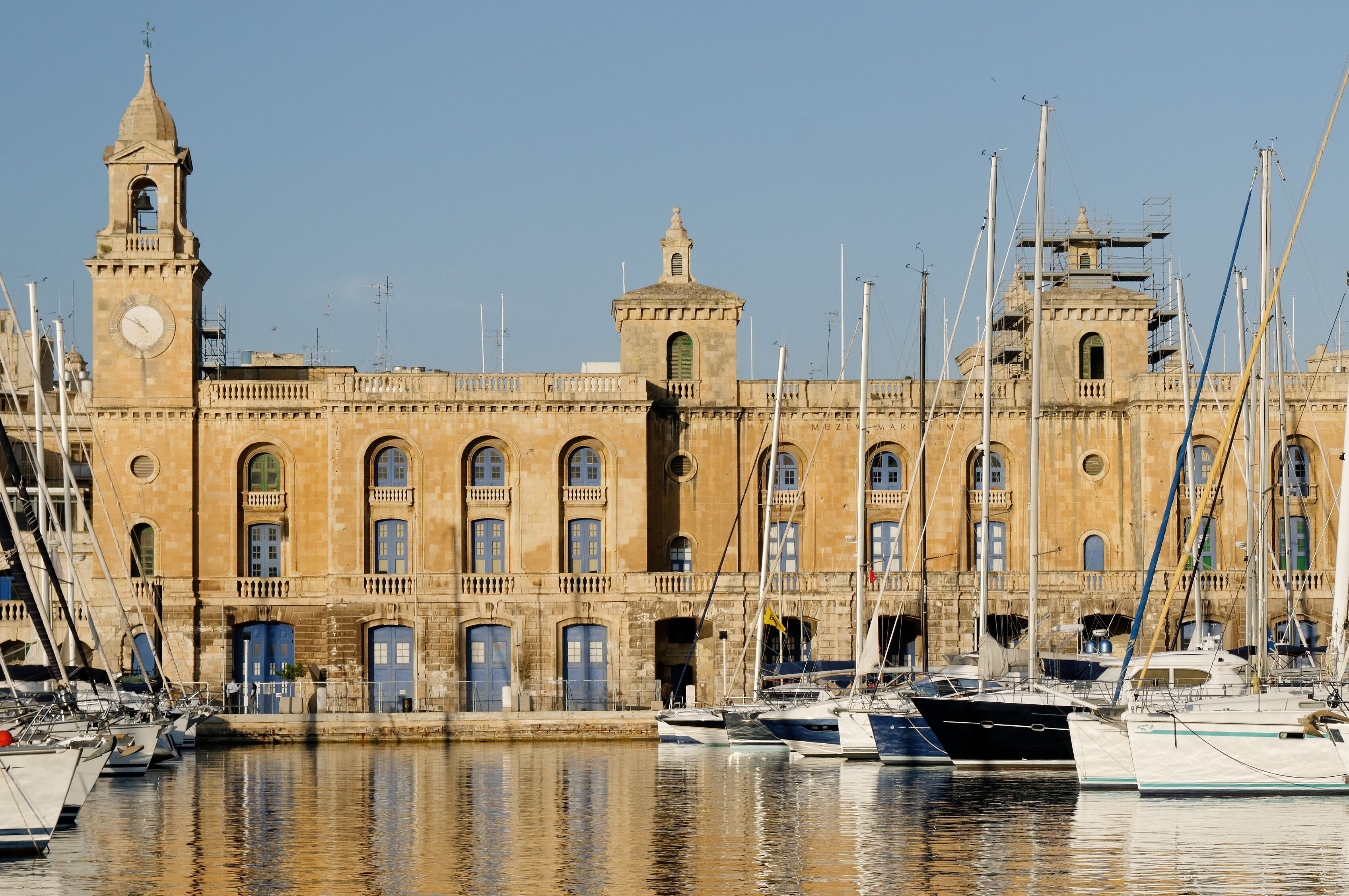 A historic building with a clock tower and a bell, surrounded by a marina with several boats.