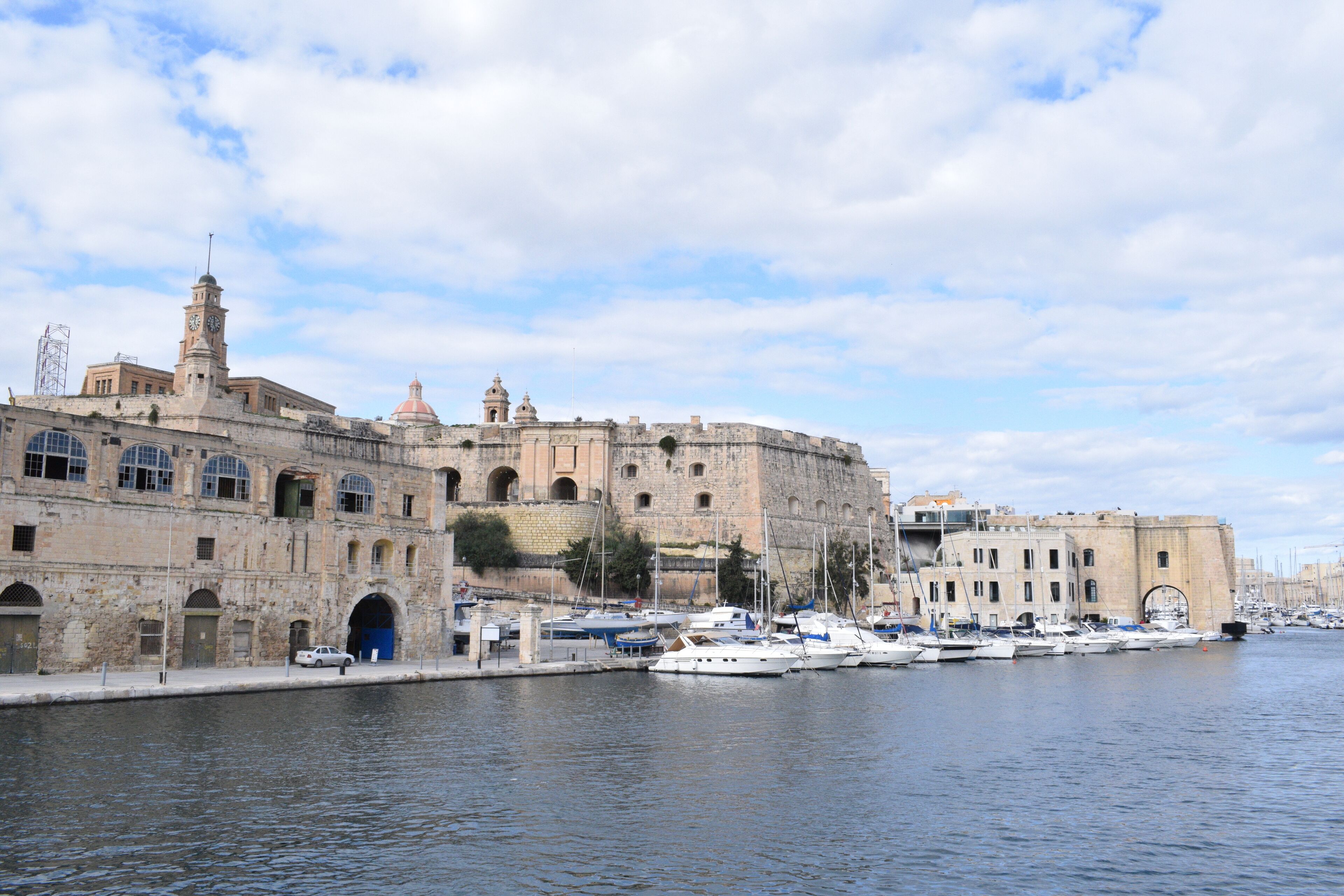 A historic stone building with a clock tower and a dome, surrounded by a marina with boats.