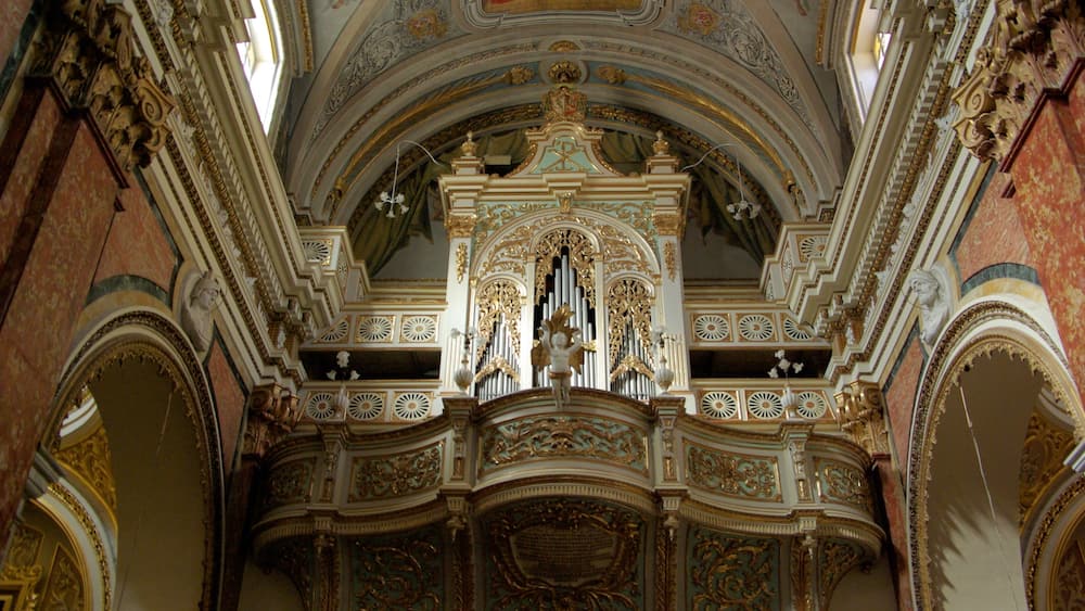 An ornate church interior with detailed gold and white decorations, a large organ, and a fresco on the ceiling.