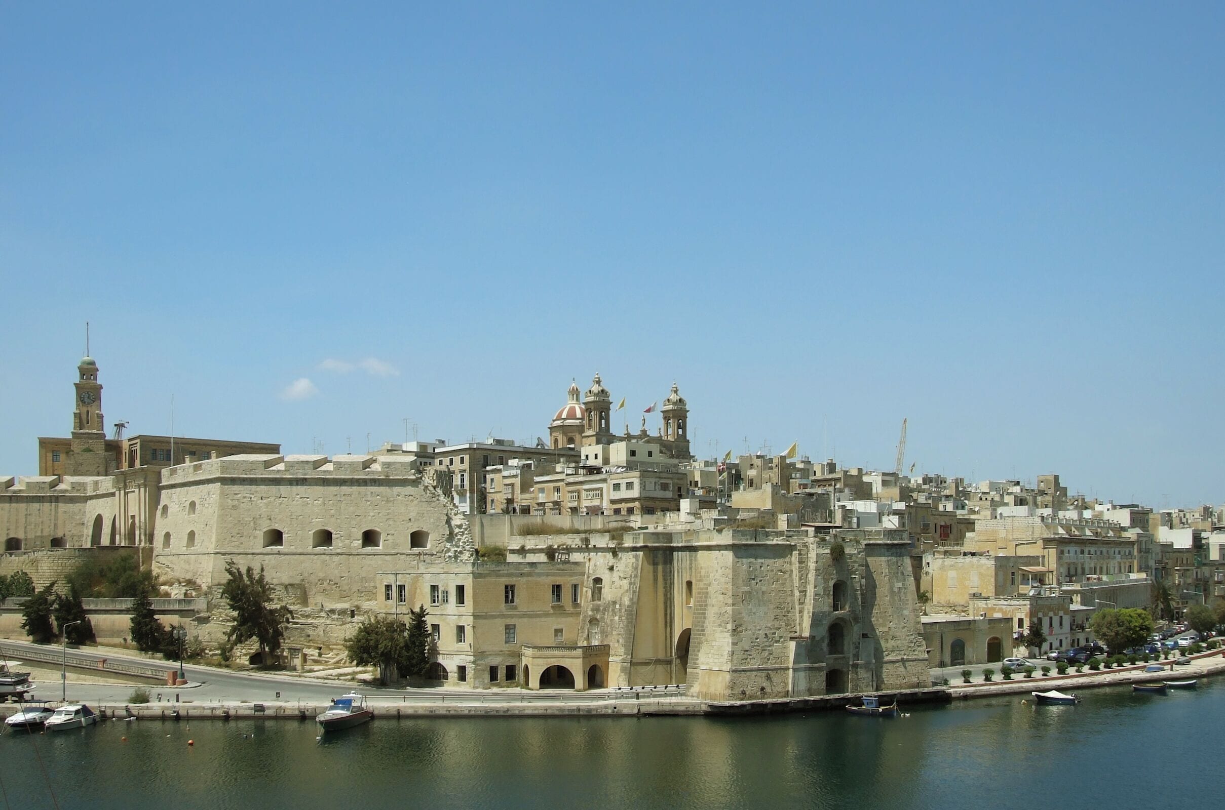 A coastal cityscape with historic buildings, a prominent clock tower, and a marina with boats.