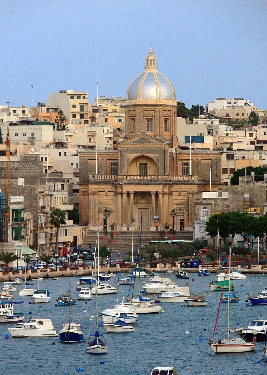 A coastal cityscape with a prominent domed building and numerous boats in the harbor.