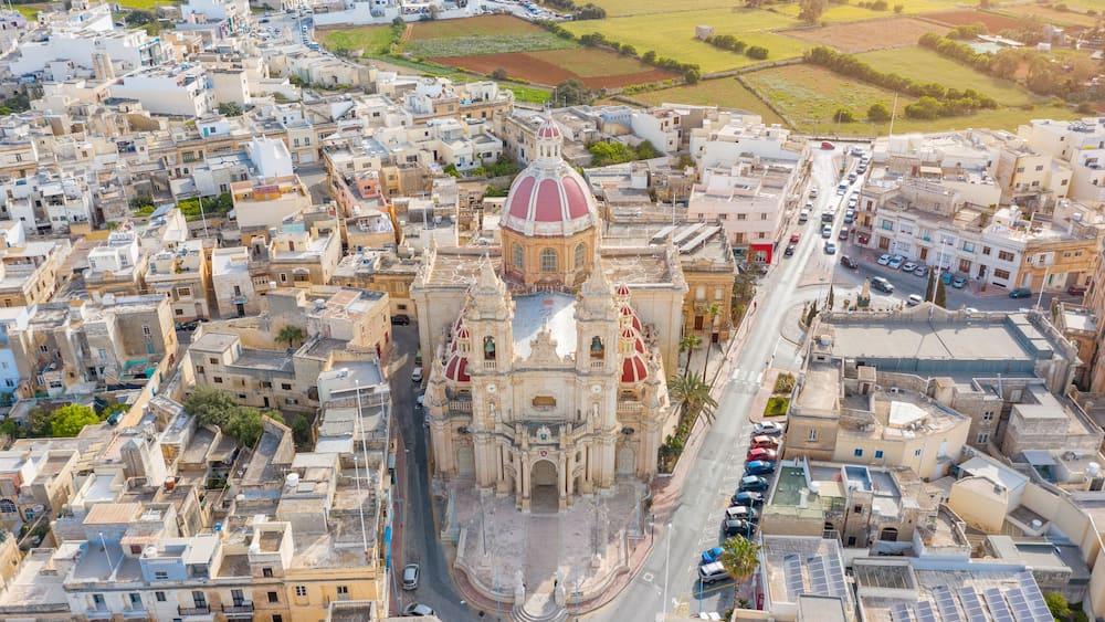 Une vue aérienne d’une ville où l’on aperçoit une église imposante, reconnaissable à son dôme rouge, entourée de bâtiments très rapprochés les uns des autres et bordée d’une route où sont garées des voitures.