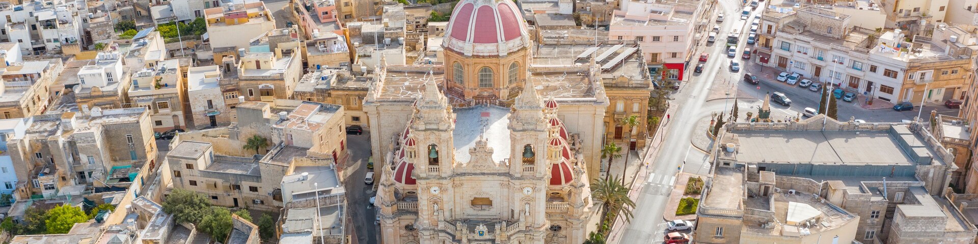 Vista aérea de una ciudad con una iglesia destacada que tiene una cúpula roja, rodeada de edificios muy cercanos y una calle con coches estacionados.