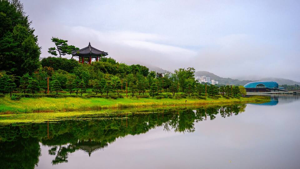 A serene lake with a traditional pavilion, lush greenery, and distant modern buildings.