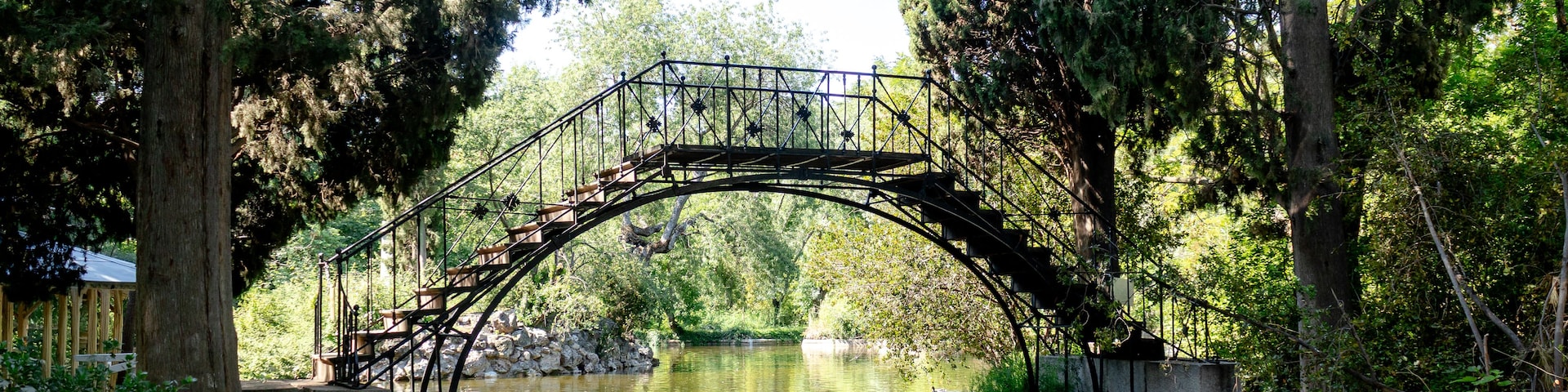 Uma ponte de pedra sobre um rio calmo, cercada por árvores e uma passarela de madeira.