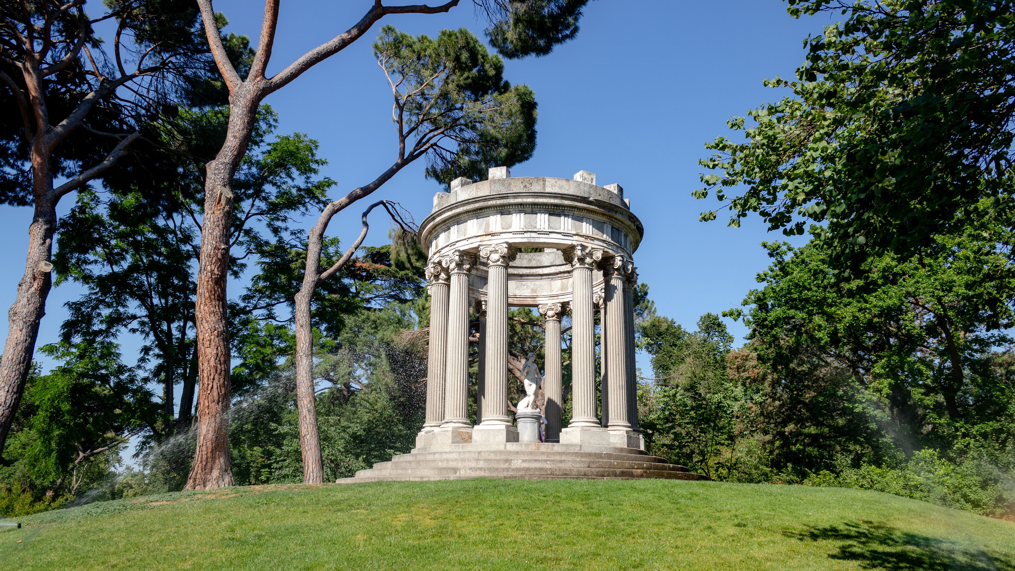 A classical-style gazebo with Corinthian columns, surrounded by lush greenery and tall trees.