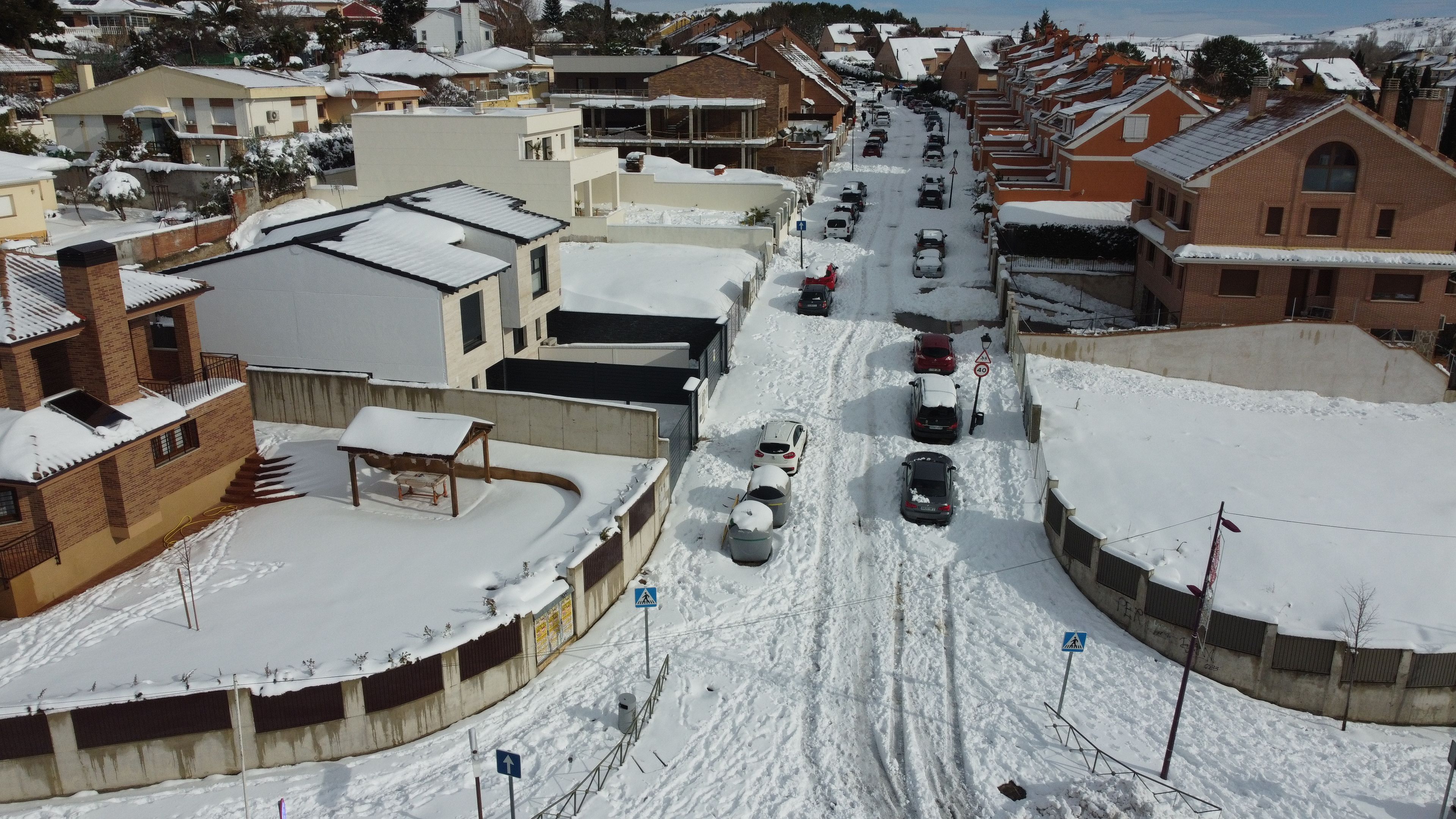 A snow-covered street with cars parked on both sides, surrounded by residential buildings.