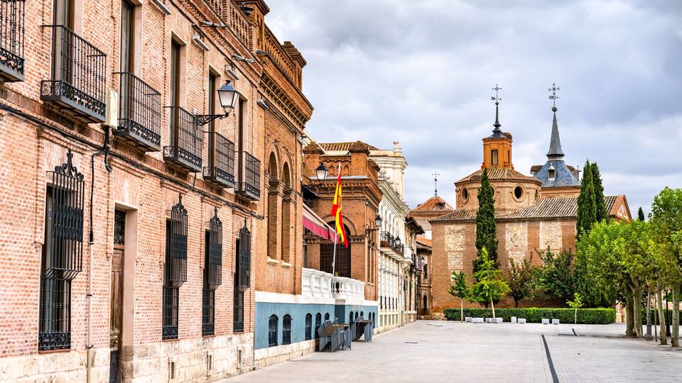 A historic plaza with a church in the background, featuring a prominent bell tower and a flagpole with a flag.