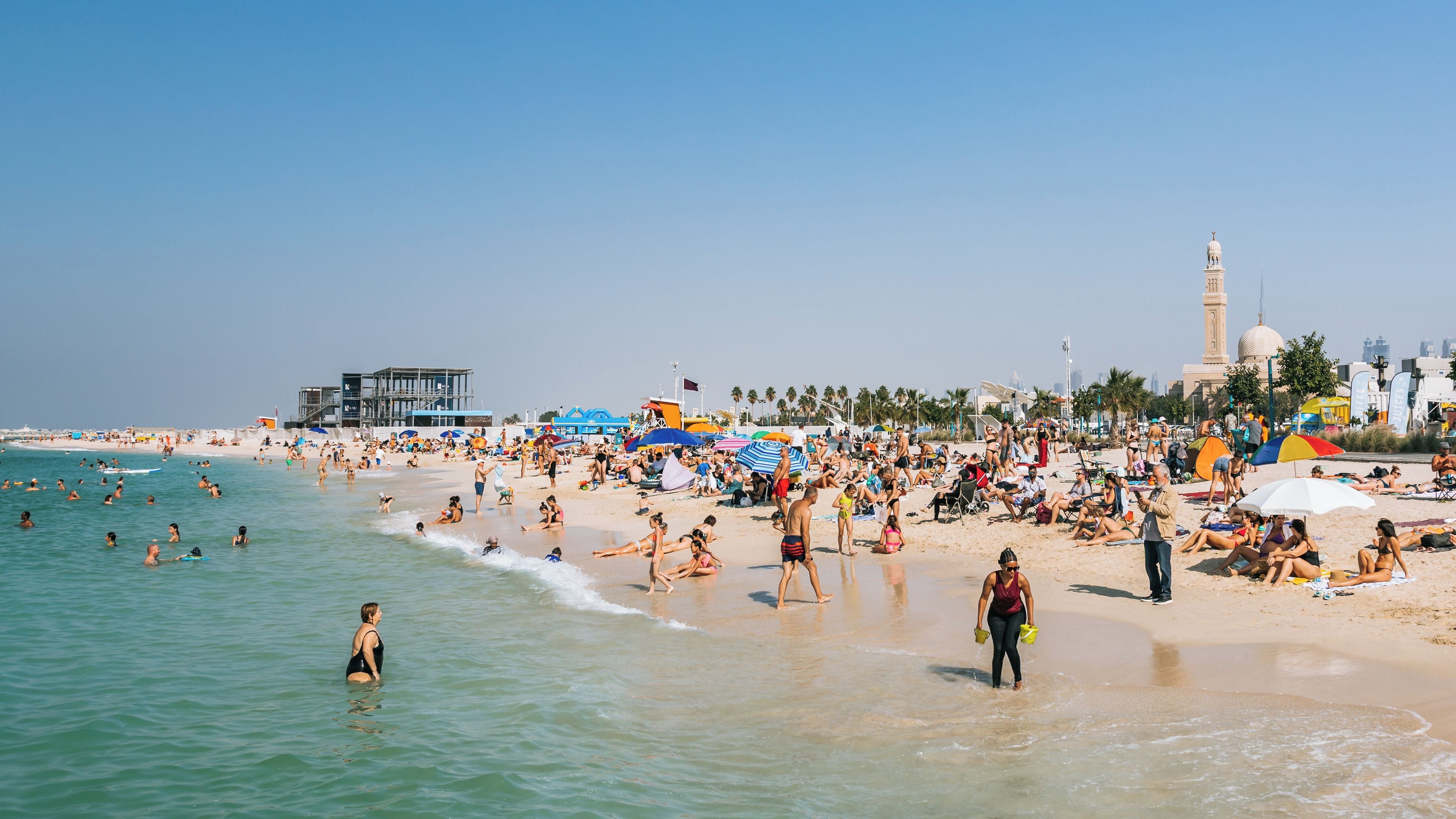 A crowded beach with people swimming and sunbathing, a lifeguard tower, and a mosque with a minaret in the background.