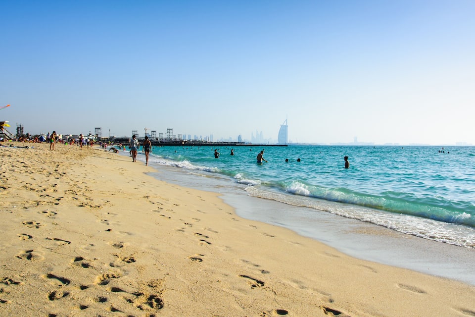 A sandy beach with footprints, calm sea, and a distant city skyline.