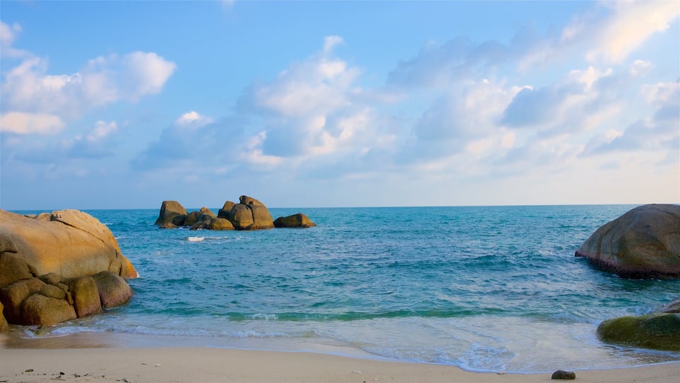 A beach with large rocks and calm blue water under a partly cloudy sky.