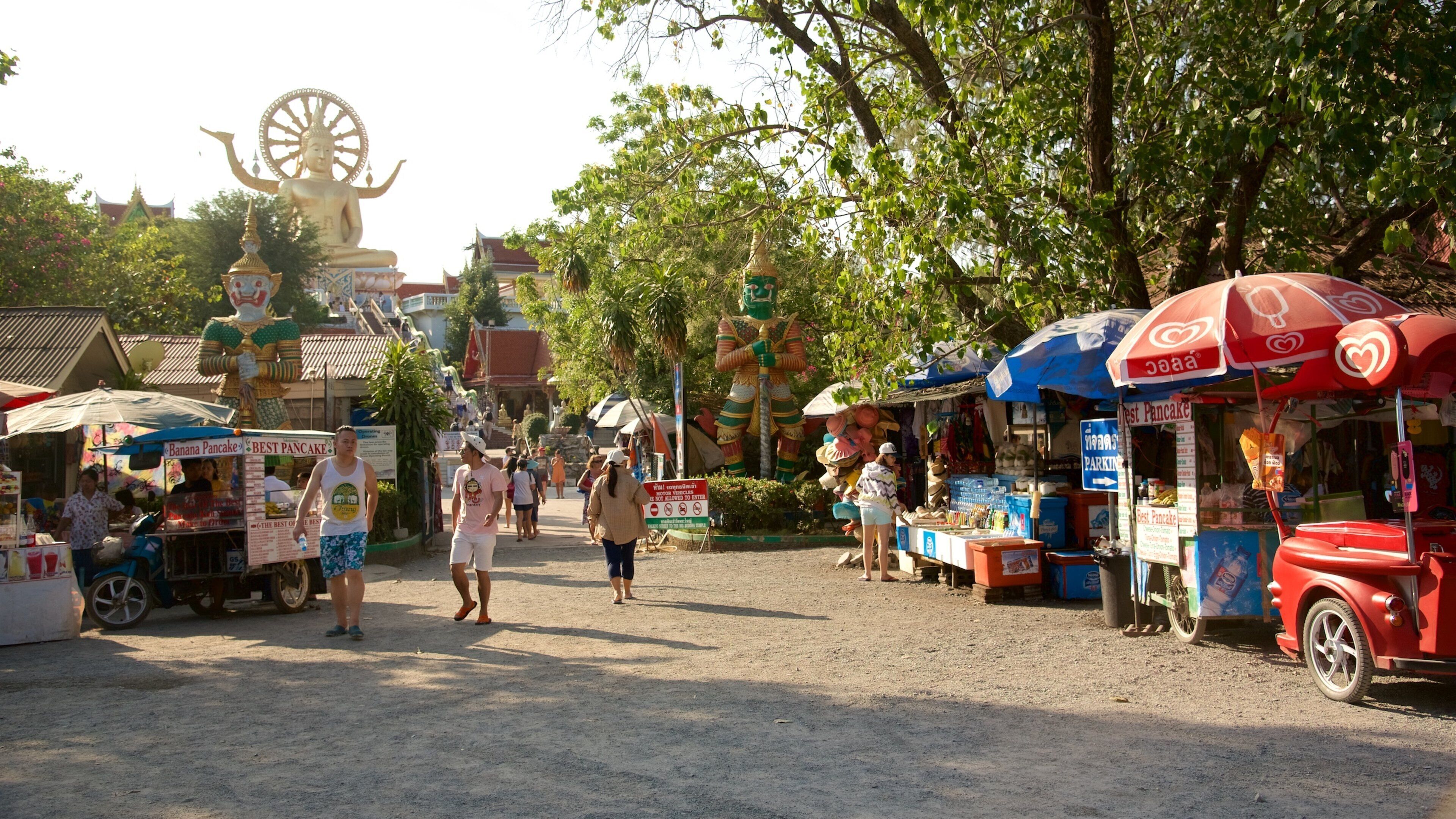 A bustling street market with food stalls, a large Buddha statue, and traditional architecture.