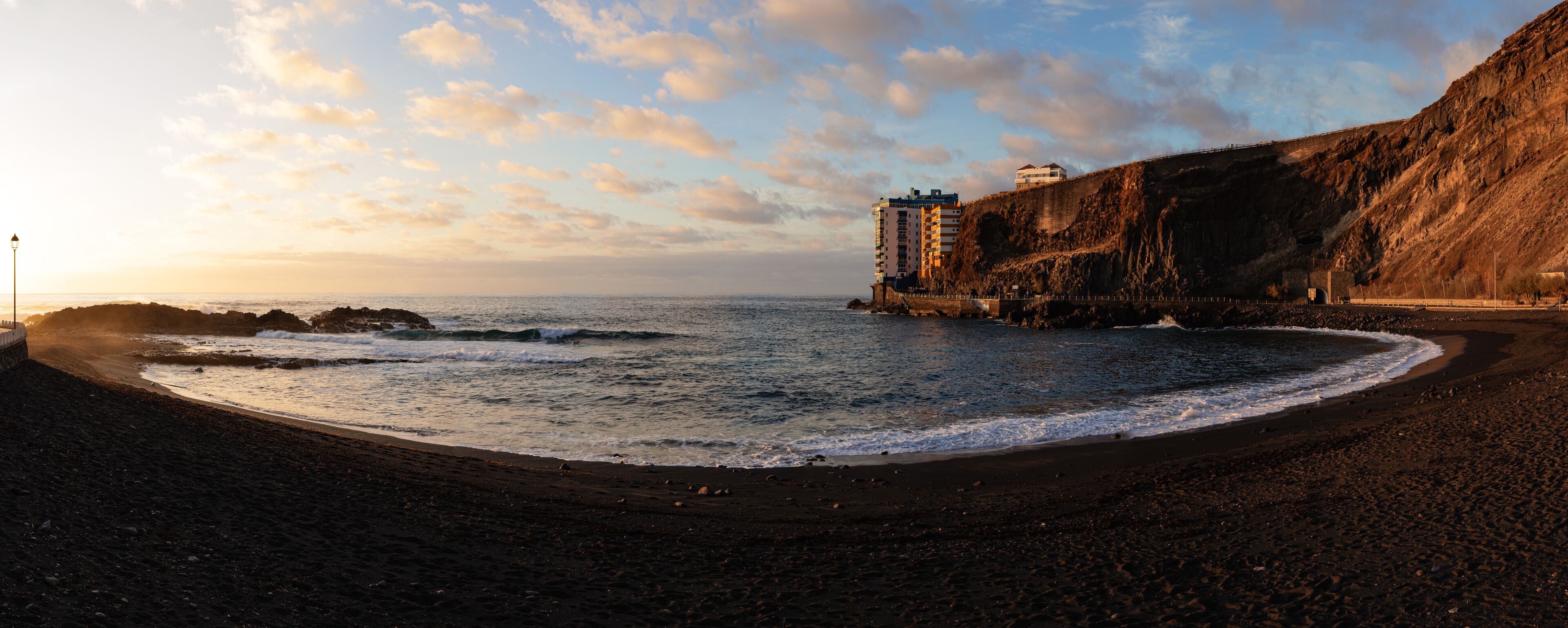 A beach with dark sand, waves, and a cliff with buildings.