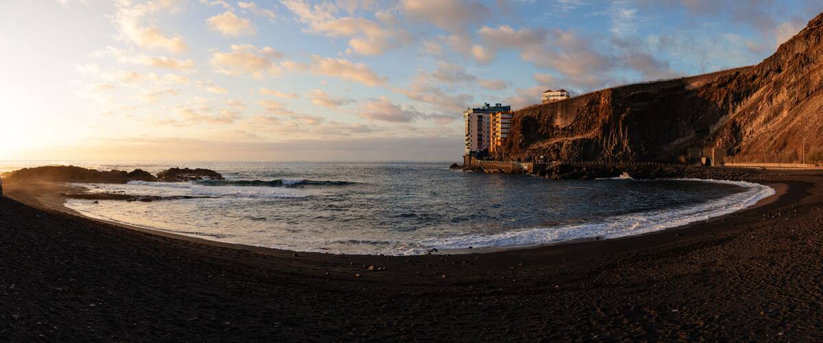 A beach with dark sand, waves, and a cliff with buildings.