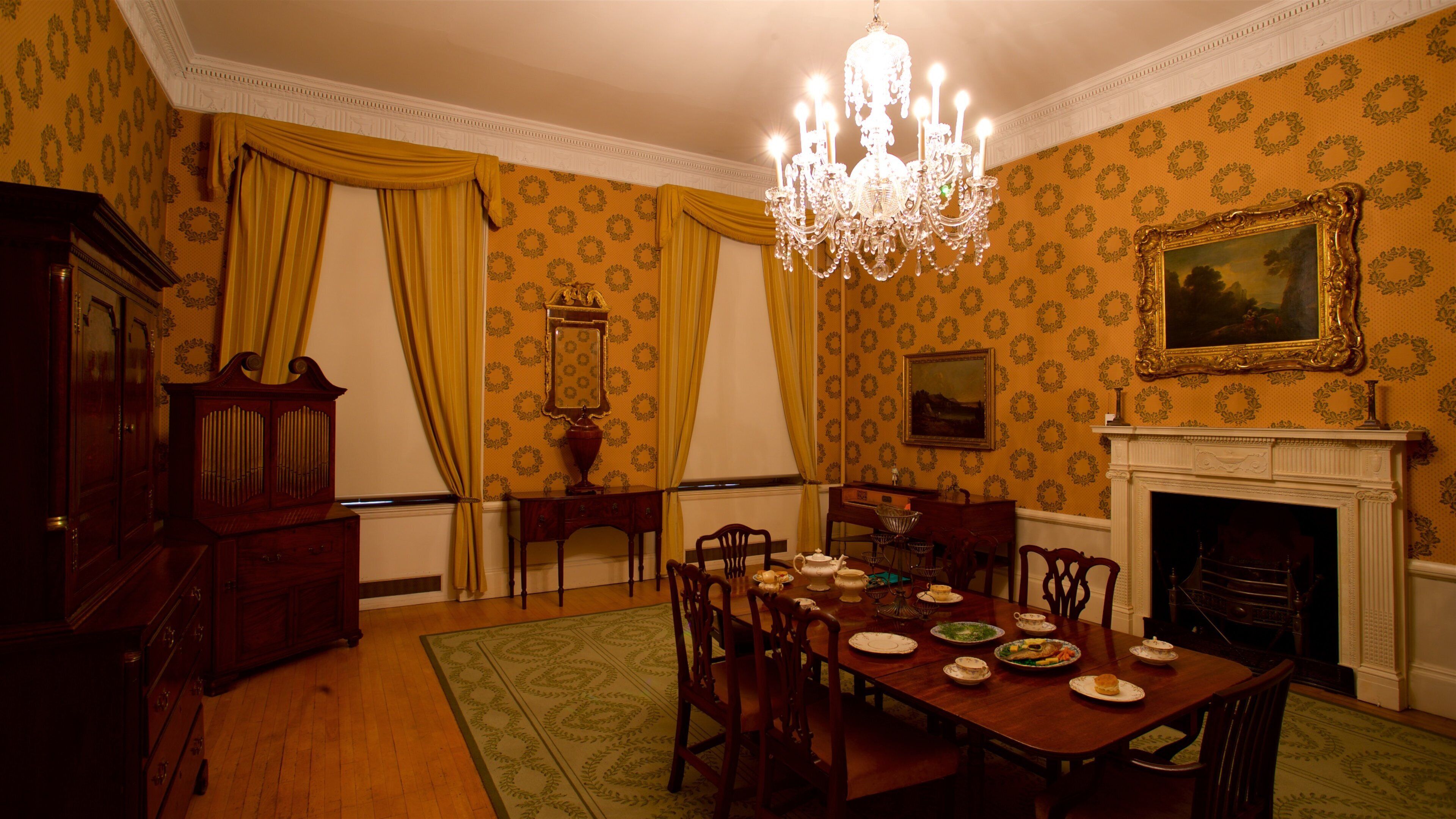 Une salle à manger avec une table en bois dressée pour le repas, un lustre, une cheminée et un papier peint à motifs.