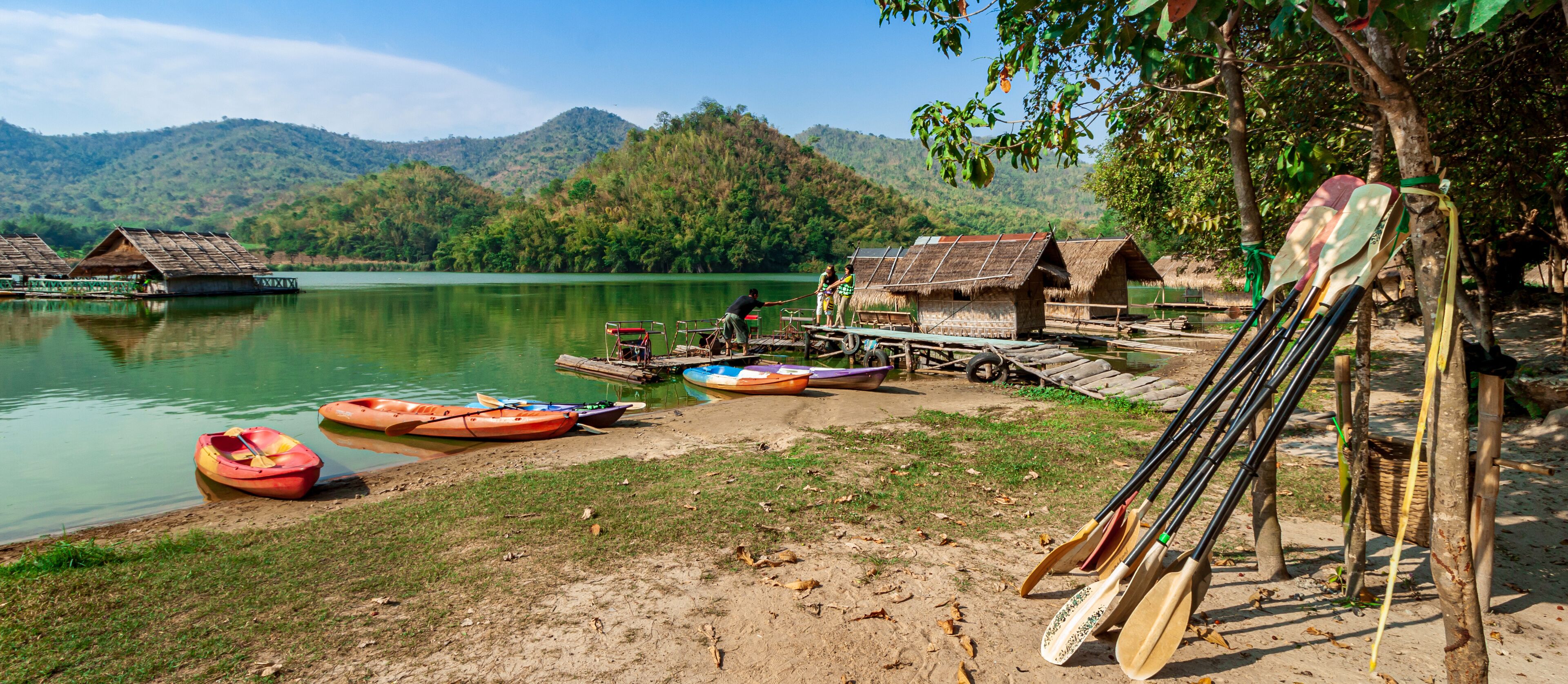 A lakeside village with thatched-roof huts, kayaks, and paddleboards by the water.