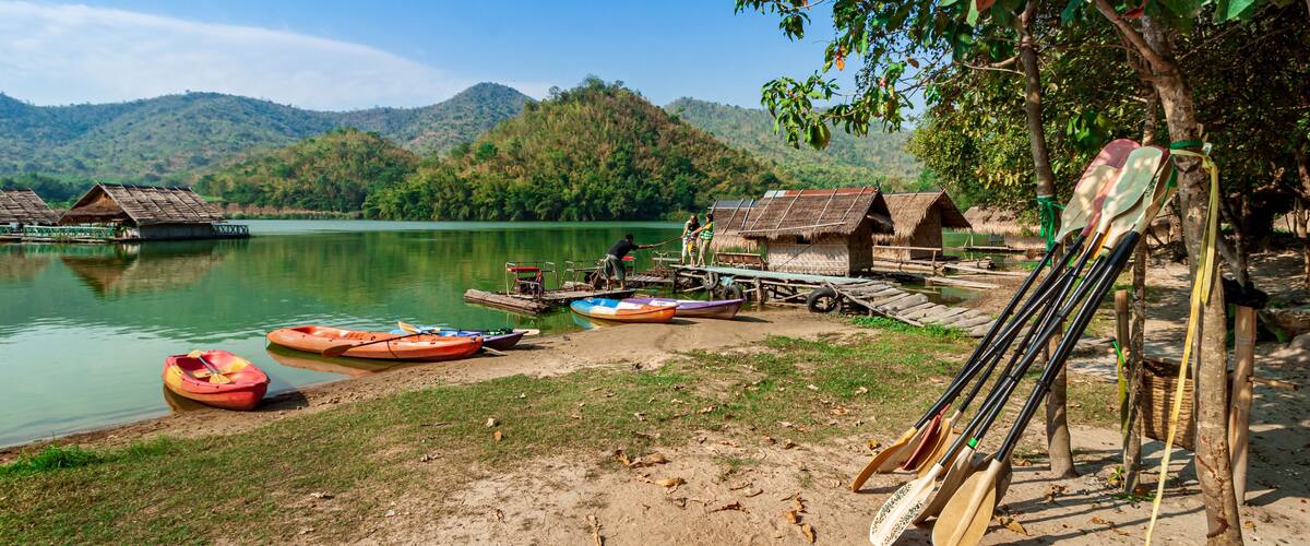 A lakeside village with thatched-roof huts, kayaks, and paddleboards by the water.