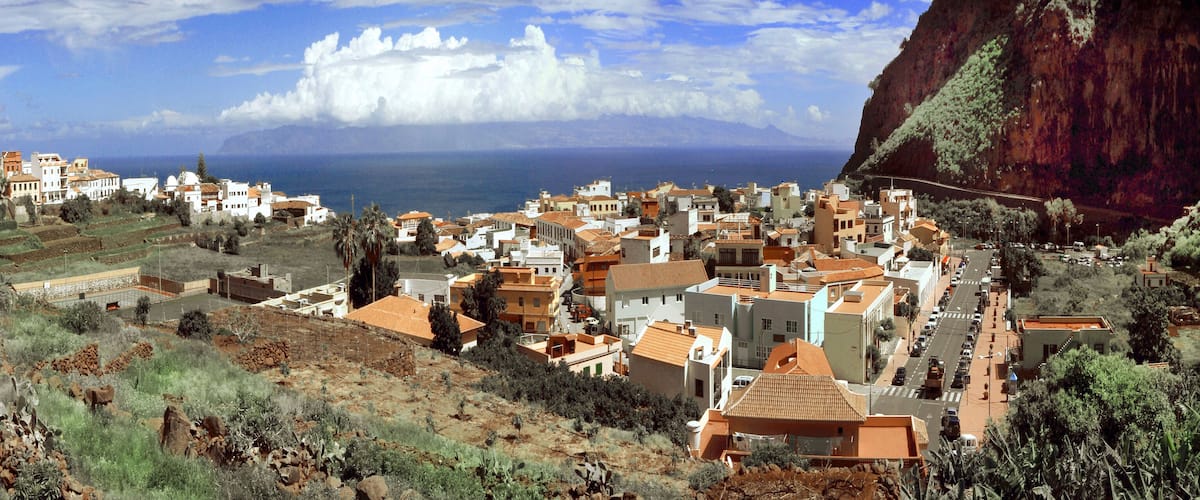 A coastal town with terraced fields, buildings with orange roofs, and a mountain in the background.