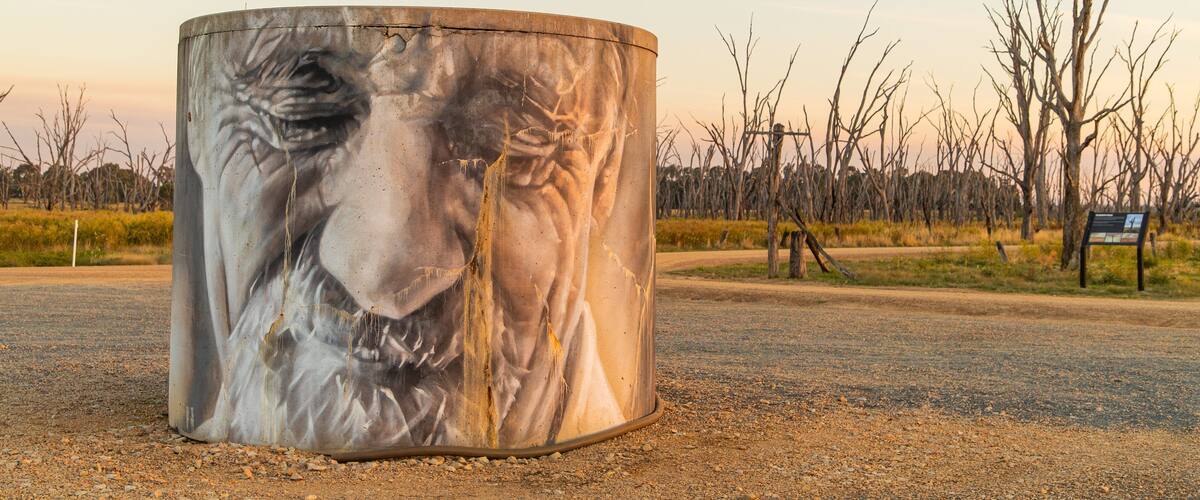 A cylindrical sculpture of a lion's face on a gravel road with leafless trees and a sign in the background.