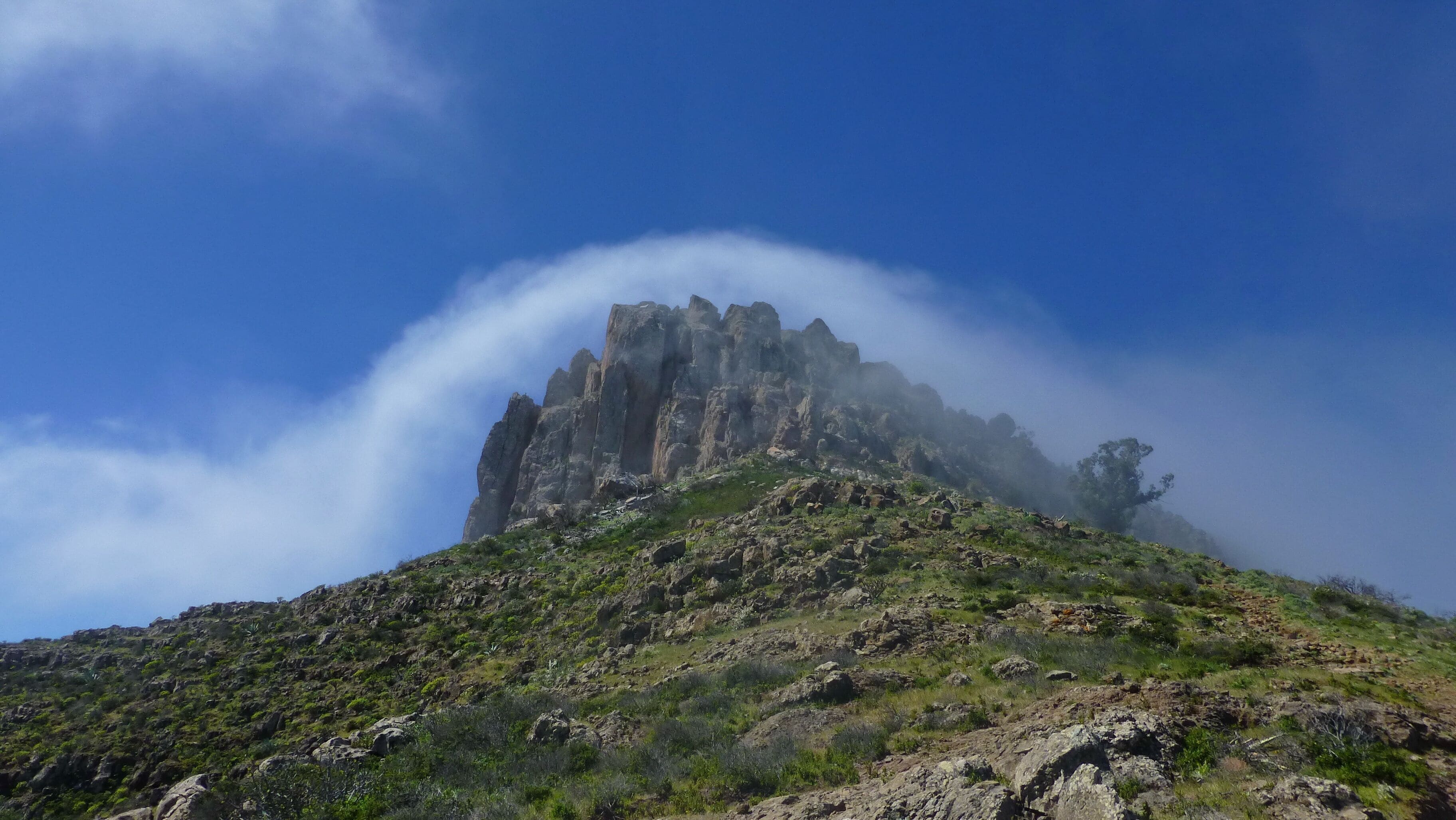 A rugged mountain peak with a prominent rock formation, surrounded by sparse vegetation and a clear blue sky.