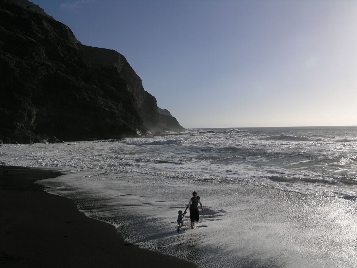 A beach with waves crashing onto the shore, a person walking with a child, and a cliff in the background.