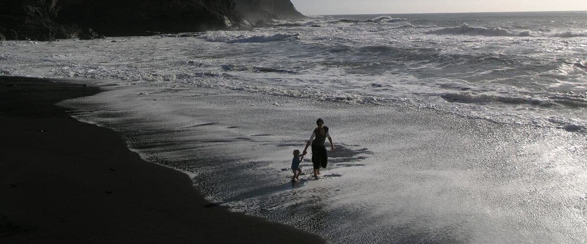 A beach with waves crashing onto the shore, a person walking with a child, and a cliff in the background.