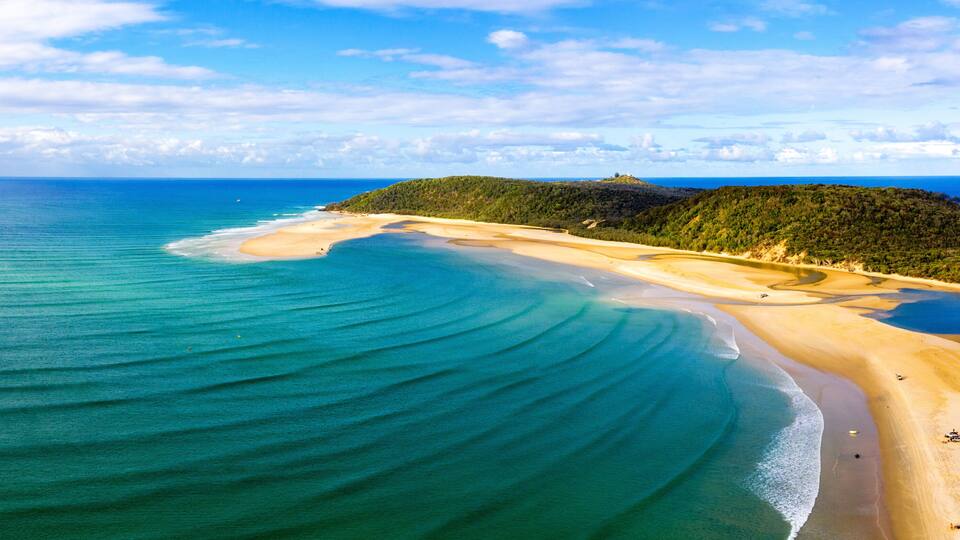 A coastal landscape with a sandy beach, clear turquoise waters, and a green hill in the distance.
