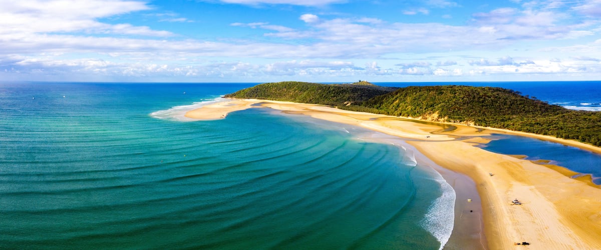A coastal landscape with a sandy beach, clear turquoise waters, and a green hill in the distance.