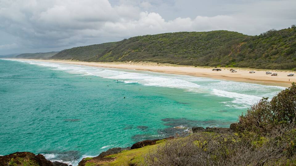 A beach with turquoise waters, a sandy shore, and a forested hill in the background.