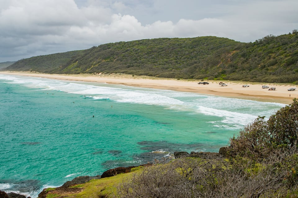 A beach with turquoise waters, a sandy shore, and a forested hill in the background.