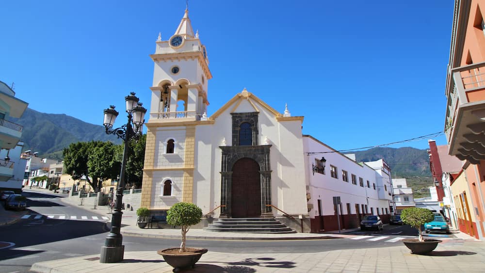 A church with a bell tower, a street with cars, and a mountain in the background.