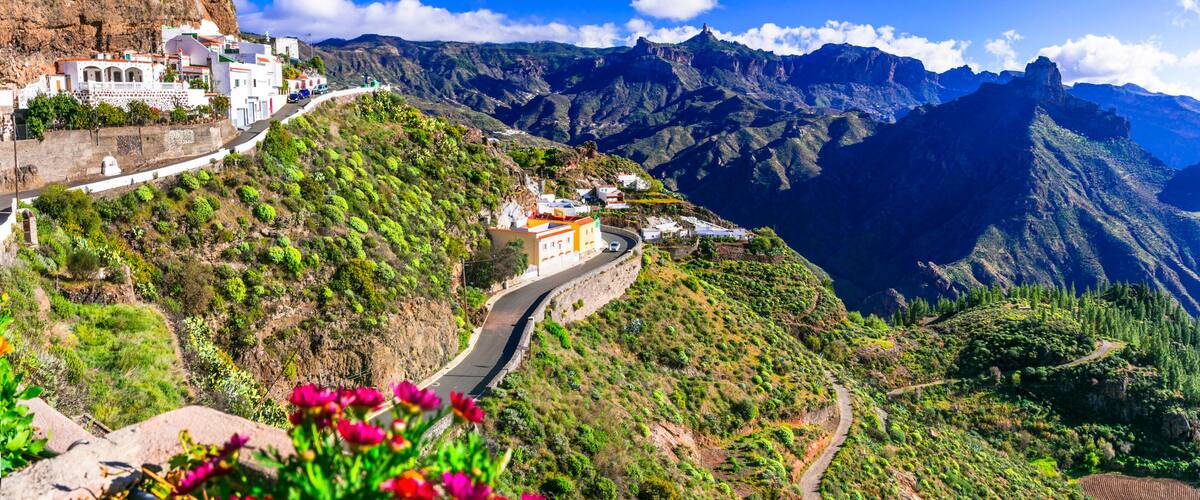 A winding mountain road with buildings on the left, surrounded by lush greenery and steep cliffs.