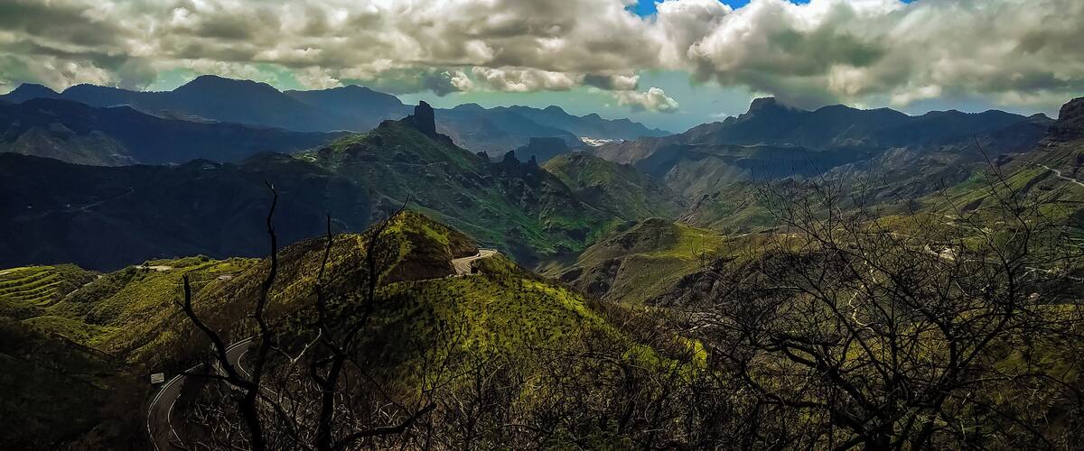 A mountainous landscape with a winding road, lush greenery, and a clear blue sky with clouds.