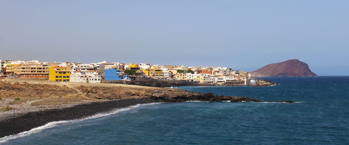 A coastal town with colorful buildings, rocky shoreline, and a prominent hill in the background.