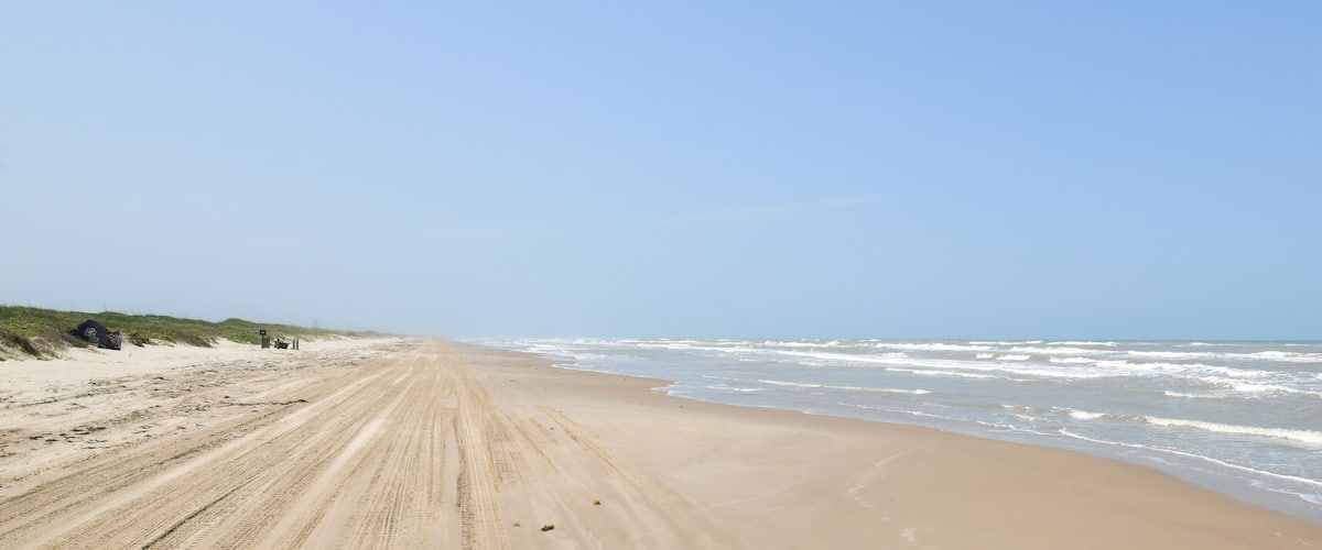 A sandy beach with waves crashing onto the shore and dunes in the background.