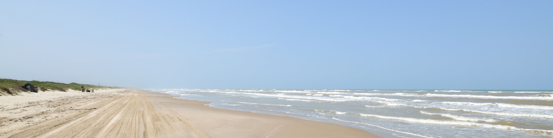 A sandy beach with waves crashing onto the shore and dunes in the background.