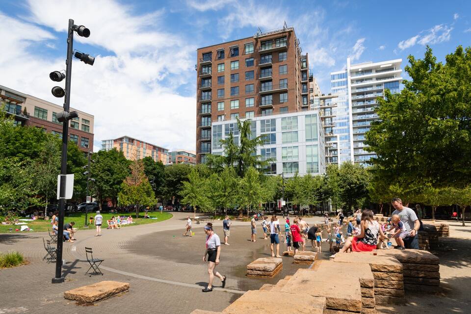 Un parque público con gente, árboles y edificios modernos al fondo.